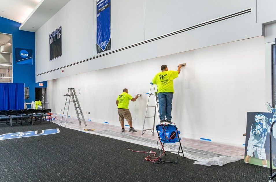 Workers painting a large white wall in a tall interior space with ladders and supplies