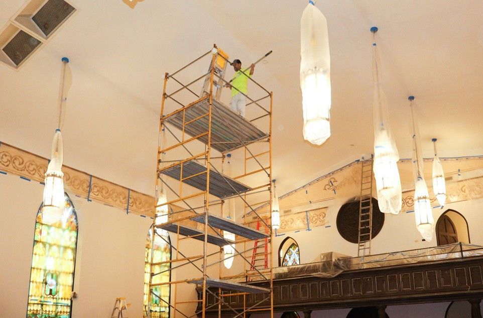 Scaffolding in a church interior with workers near hanging pendant lights and stained glass windows