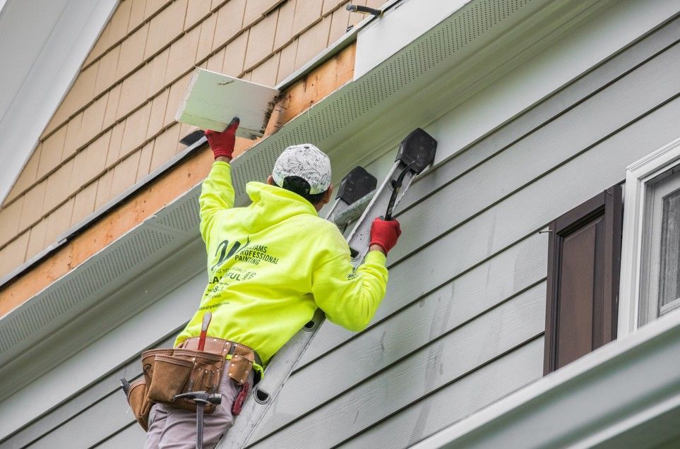 Worker in a neon jacket painting a house’s white siding on a ladder