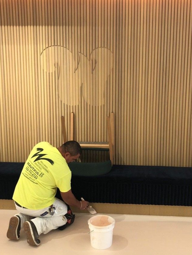 Worker kneels to paint a wood-paneled wall with a white bucket beside him.
