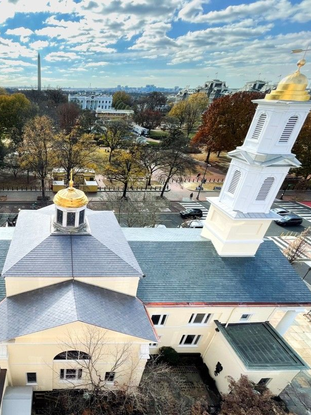 Aerial view of a white church with gold domes and steeple beside a tree-lined street under a cloudy sky