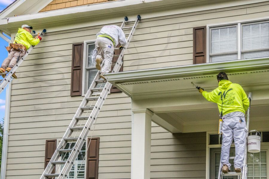 Workers painting a house exterior on ladders and scaffolding under a porch roof