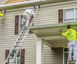 Two workers painting a house exterior from ladders, one on a roof edge and one by the porch.