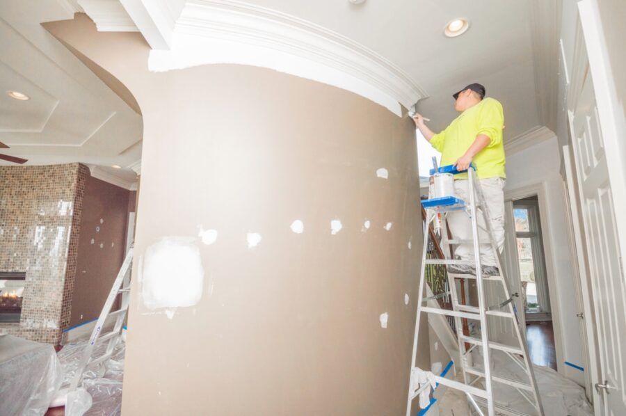 Worker painting a large curved wall inside a bright, unfinished hallway on a ladder