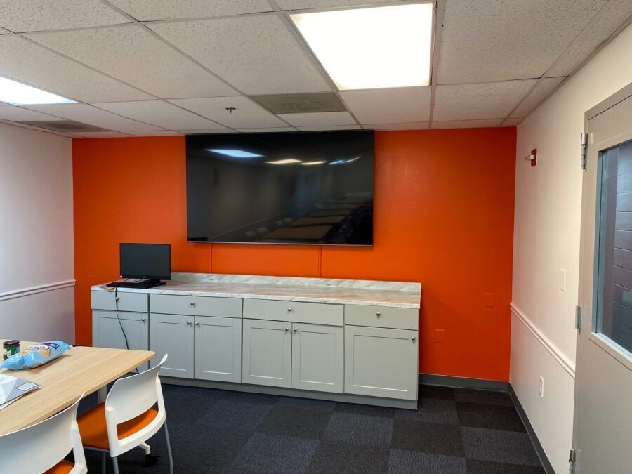 Conference room with orange accent wall, large wall-mounted TV, white cabinets, and a table in the foreground
