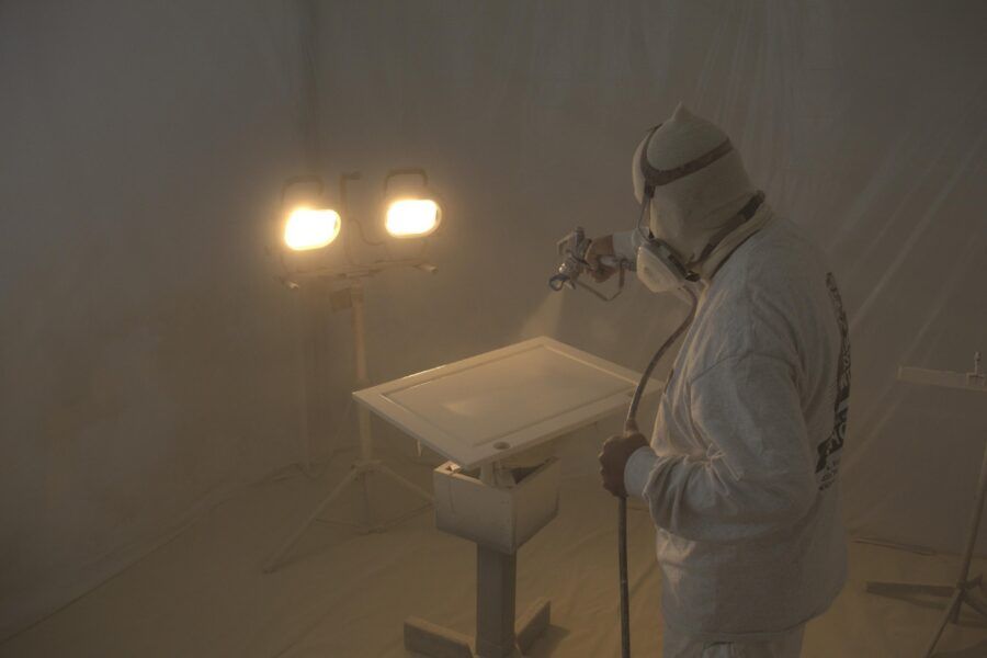 Person in white protective suit spraying a work surface in a dusty, dimly lit room.