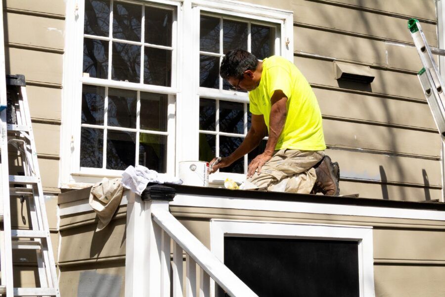 Worker in a neon yellow shirt painting a house exterior beside a window.