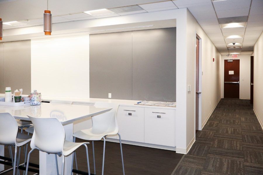 Office break room with white tables, cabinets, and chairs beside a hallway