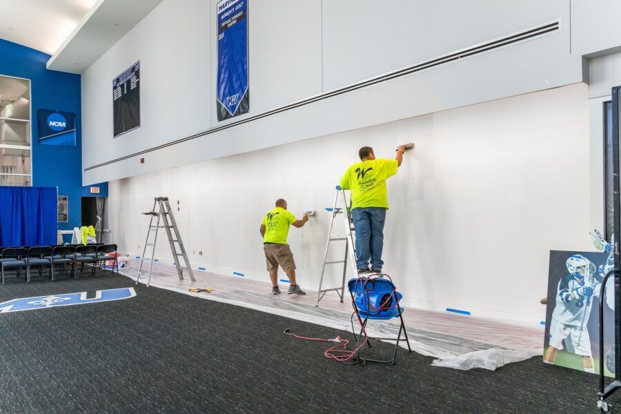 Workers painting a large white wall inside a gym, with ladders, drop cloths, and equipment nearby.