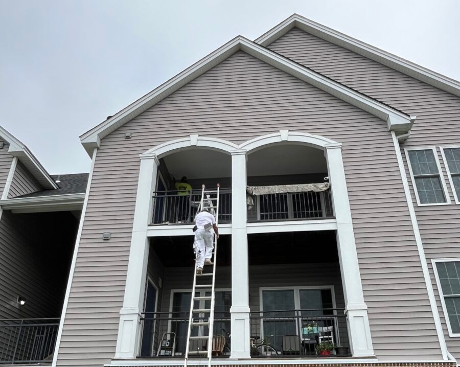 Person on ladder cleaning upper balcony of beige townhouse exterior