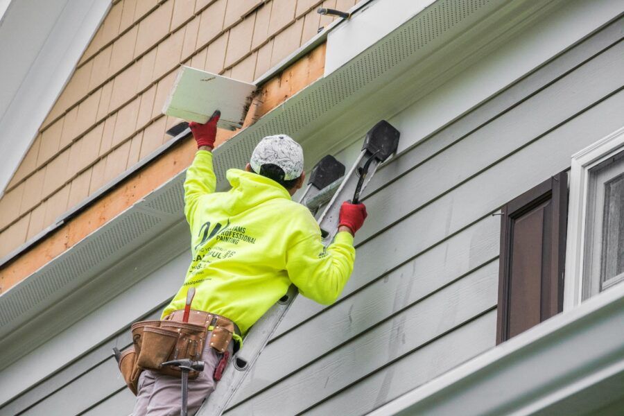 Worker in a neon jacket painting a house’s gray exterior with a roller on a ladder.