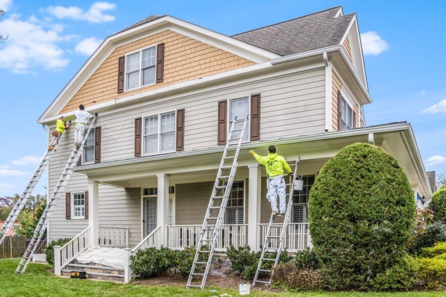 Workers painting a beige two-story house with ladders and scaffolding on the porch