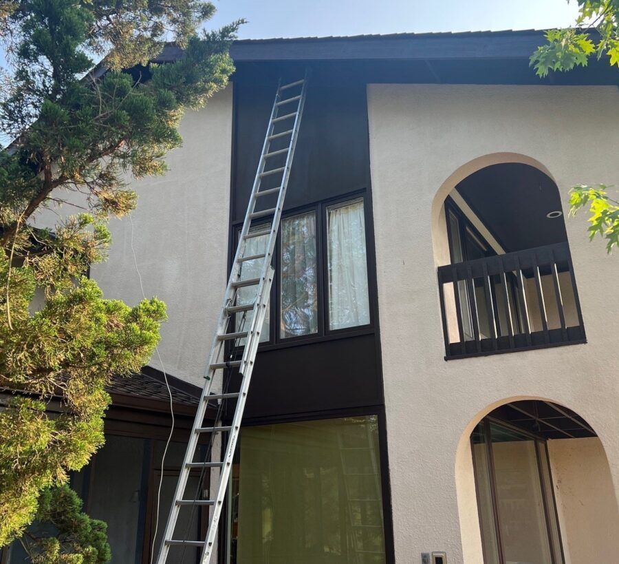 Ladder leaning against a two-story house exterior beside a window and arched balcony.