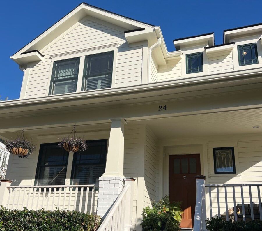 Two-story cream house with front porch, brown door, black railings, and hanging flower baskets under blue sky