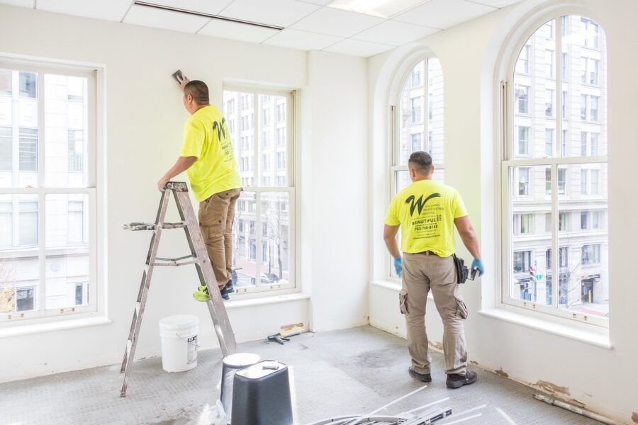 Two workers painting a bright, empty room with tall windows and a ladder