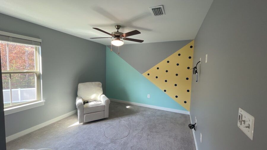 Empty gray bedroom with a white armchair, ceiling fan, and a blue-yellow geometric accent wall.