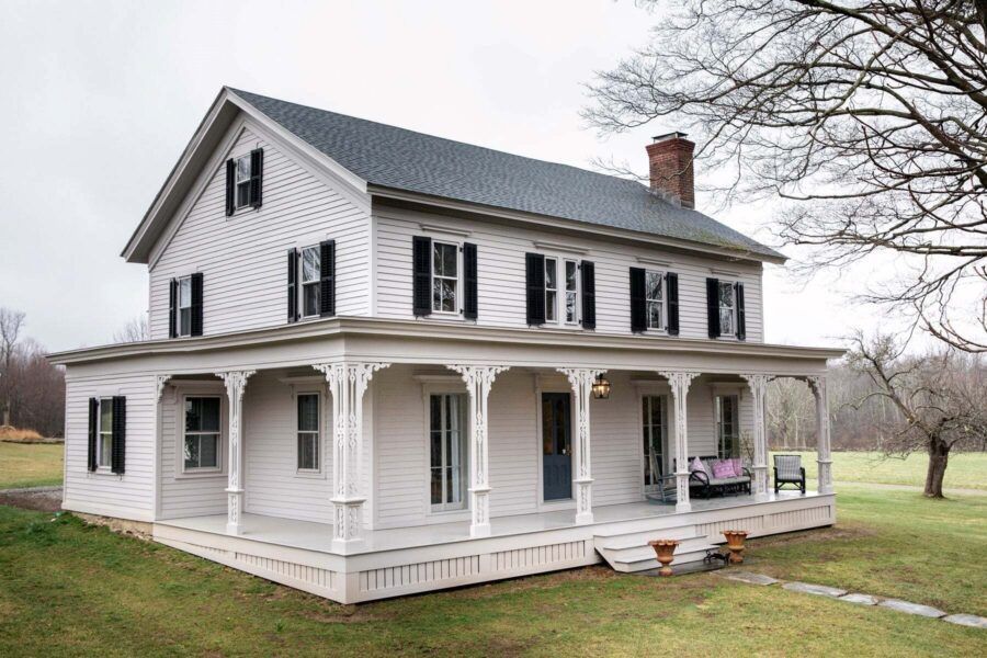 White two-story farmhouse with a wraparound porch on a grassy rural lot