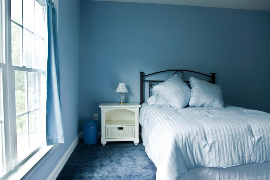 Bright blue bedroom with white bed, nightstand, lamp, and large window on the left.