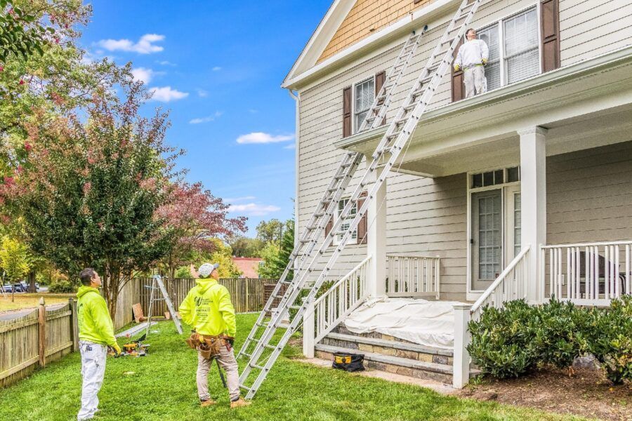 Two workers setting up an extension ladder beside a two-story house with white siding and a porch.