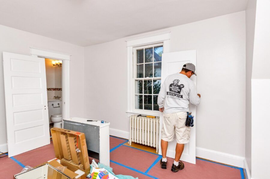 Person painting a white room with blue tape on the floor and cardboard covering furniture.