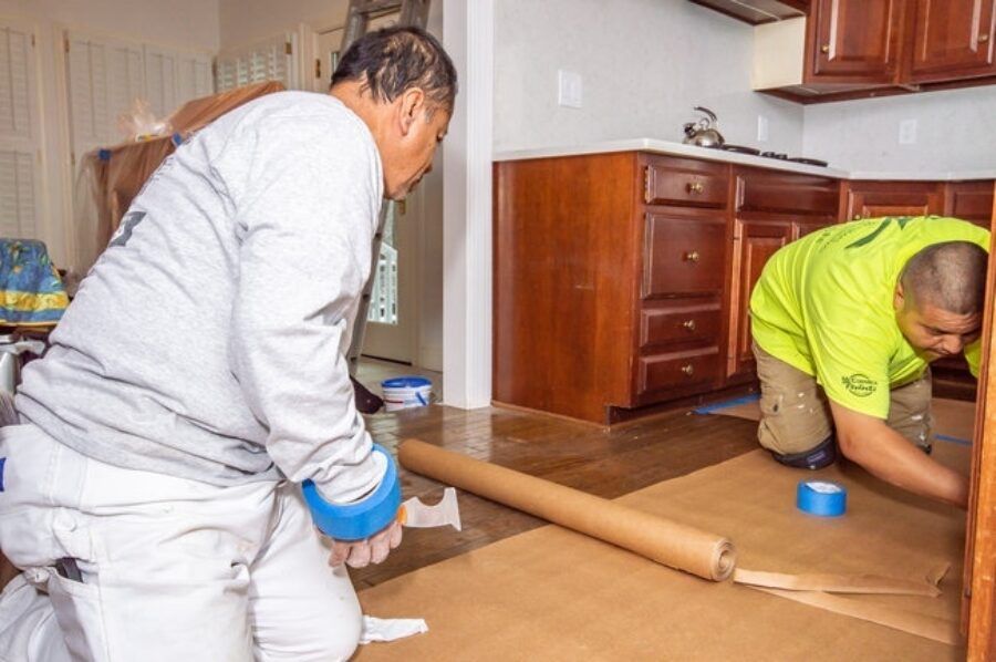 Two people unrolling flooring in a kitchen, working near wooden cabinets and a brown floor.