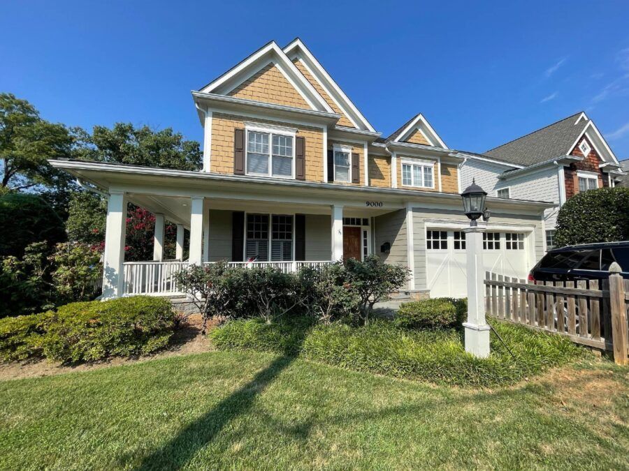 Two-story yellow house with white porch, green lawn, and wooden fence under a blue sky