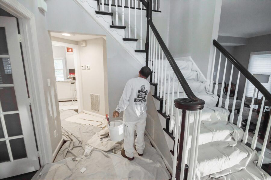Worker in white coveralls clearing debris in a damaged hallway with stairs and torn plastic sheeting.