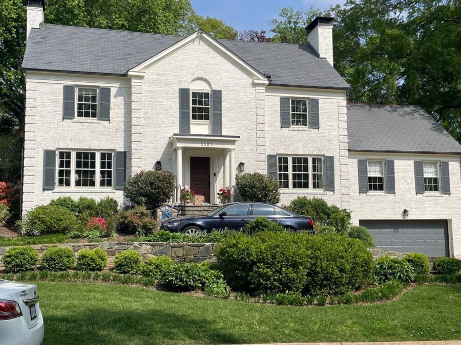 Large white two-story house with black shutters, landscaped front yard, and cars parked in the driveway
