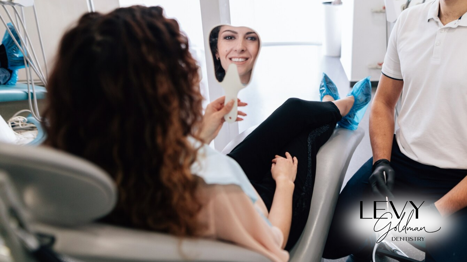 A woman is sitting in a dental chair looking at her smile in a mirror.