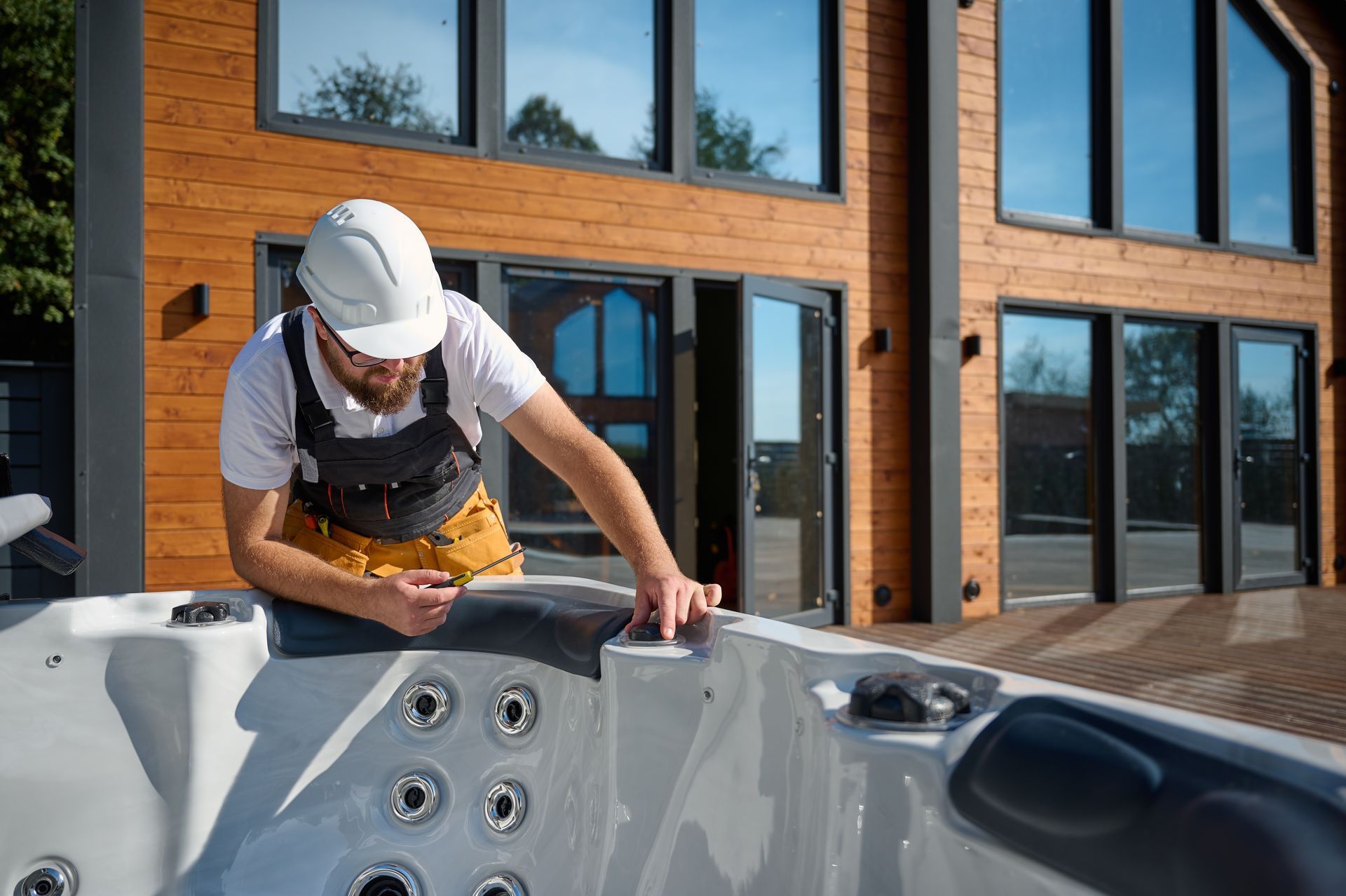 Worker wearing a hard hat installs a fixture on a rooftop spa beside a modern brick building.