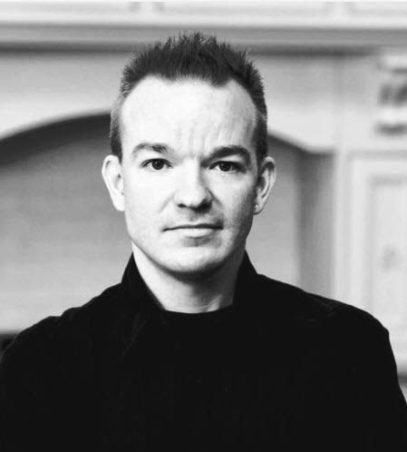 A man in a black jacket is standing in front of a fireplace in a black and white photo.