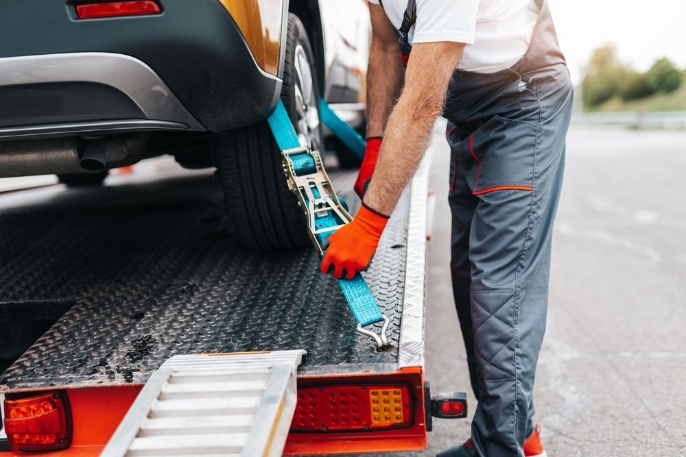 A tow truck operator secures a car to a flatbed with blue straps, wearing orange gloves and gray overalls.