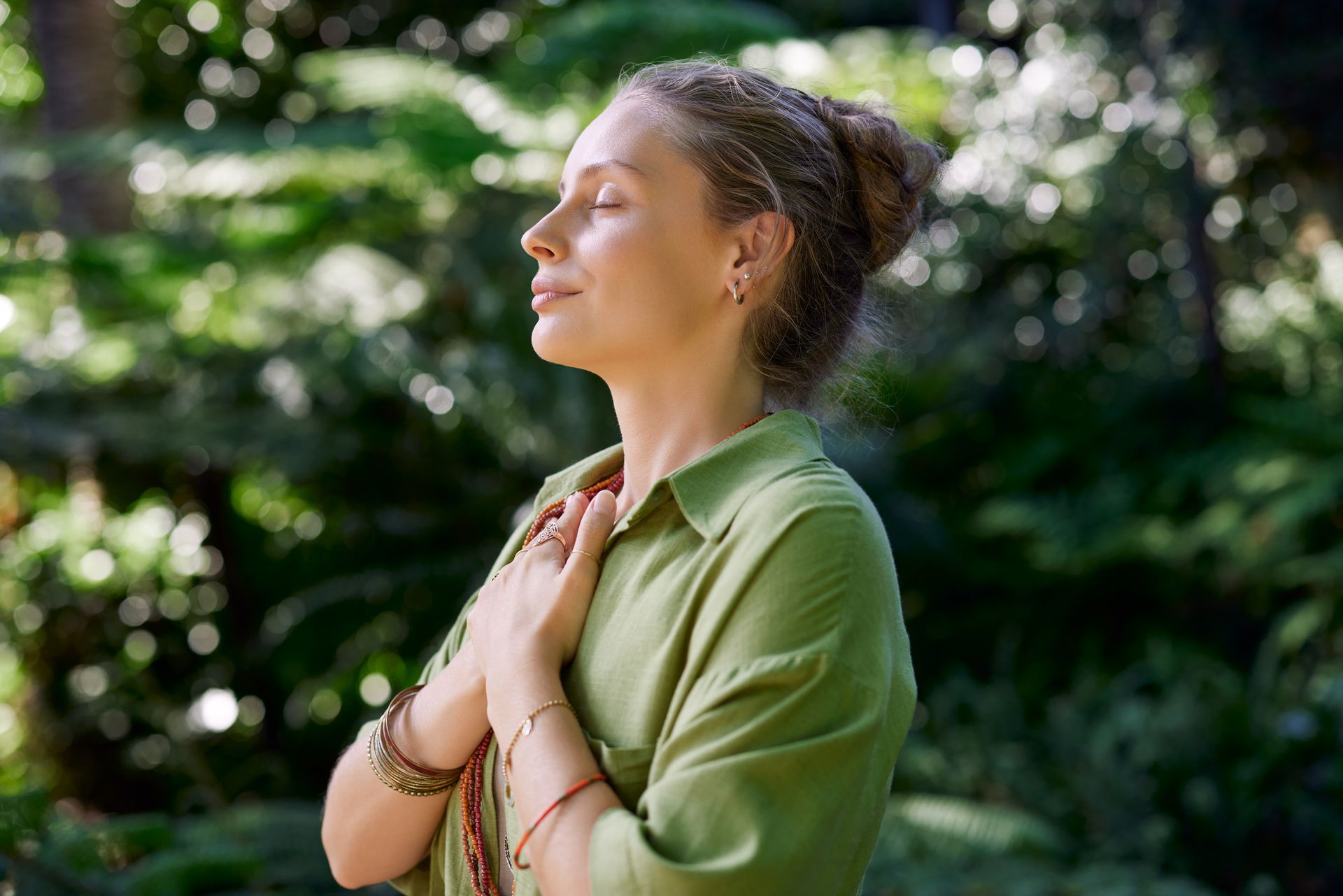 Woman in a green shirt stands in profile with eyes closed in a leafy outdoor setting. Person in a green shirt stands with hands on chest in a sunlit, leafy outdoor setting.
