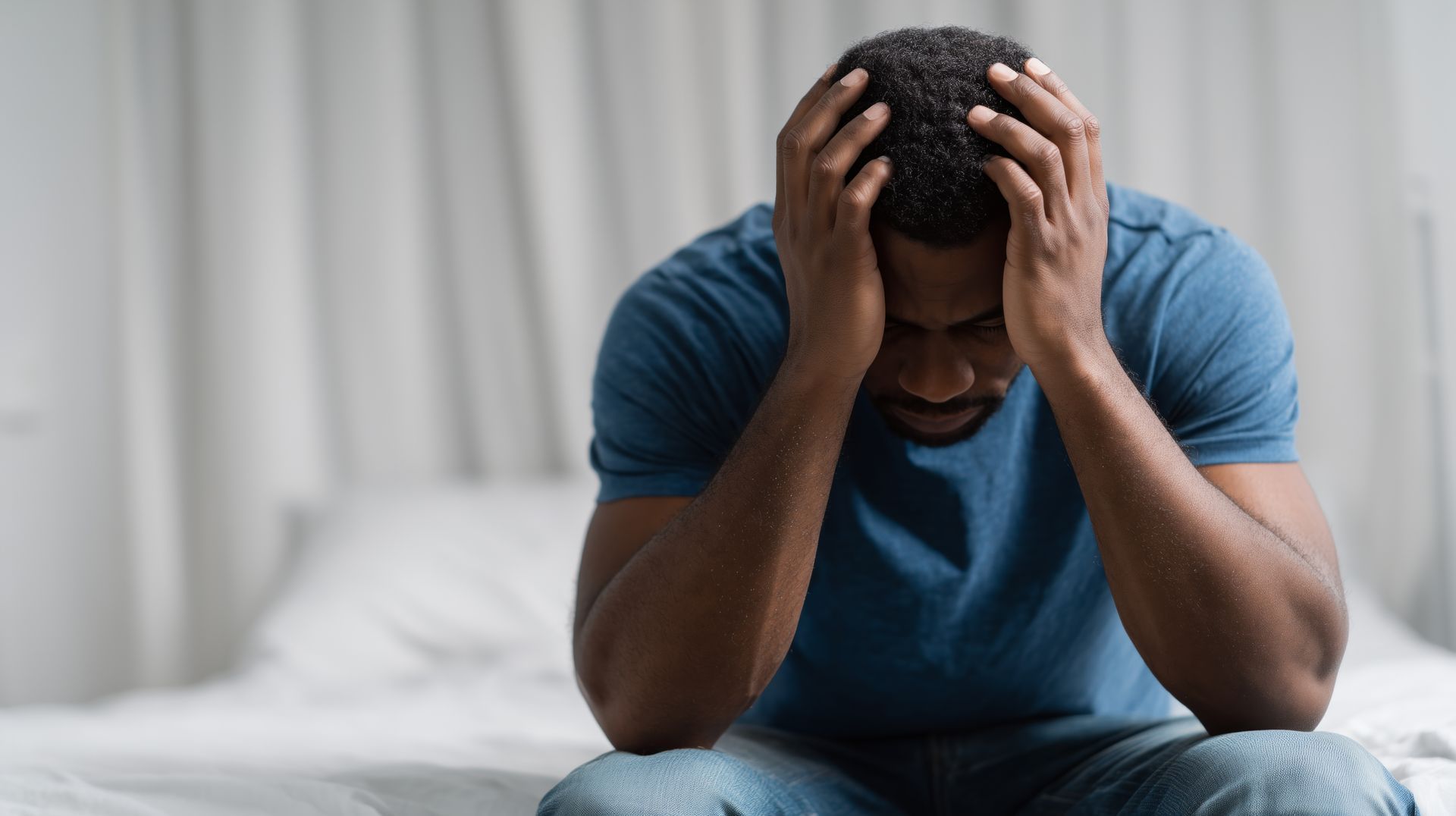 Man in a blue shirt sitting on a bed with his head in his hands, appearing stressed or distressed