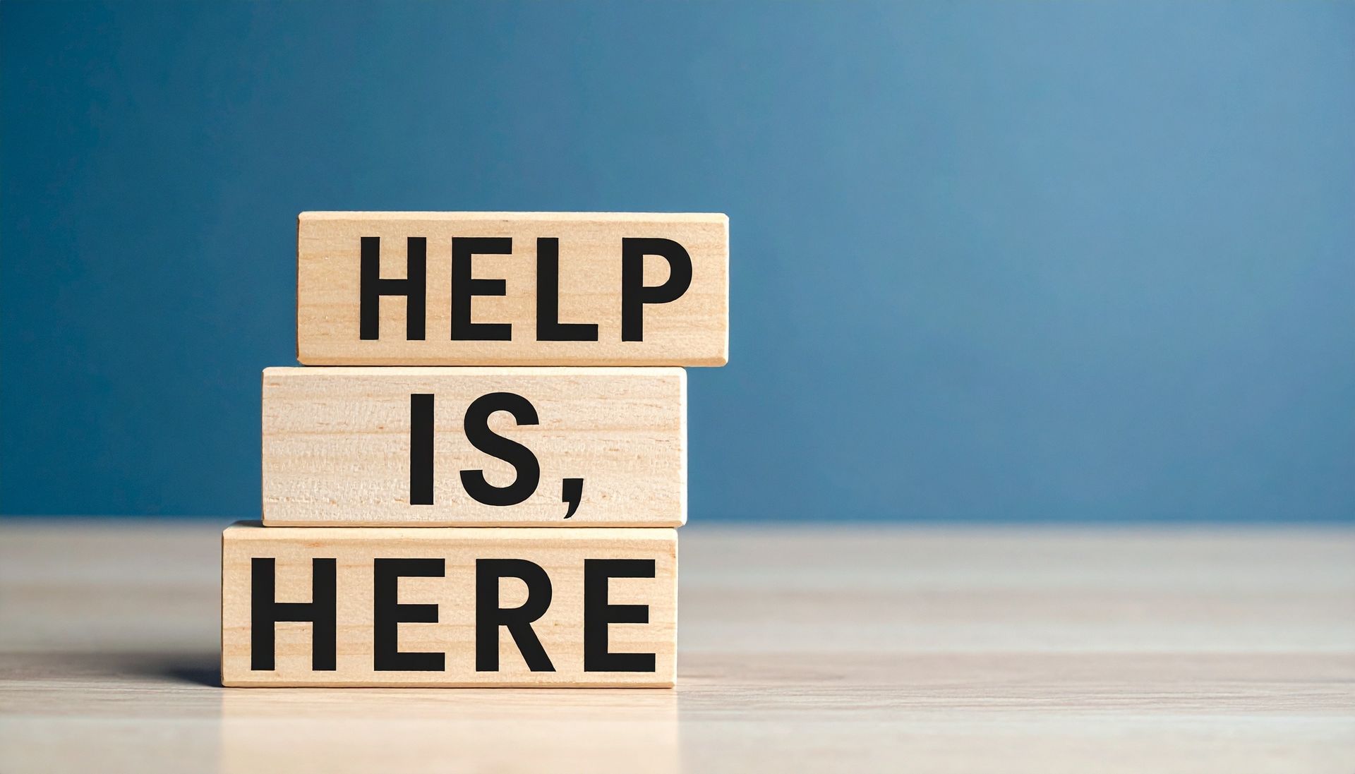 Wooden blocks spelling “HELP IS HERE” on a table against a blue background.