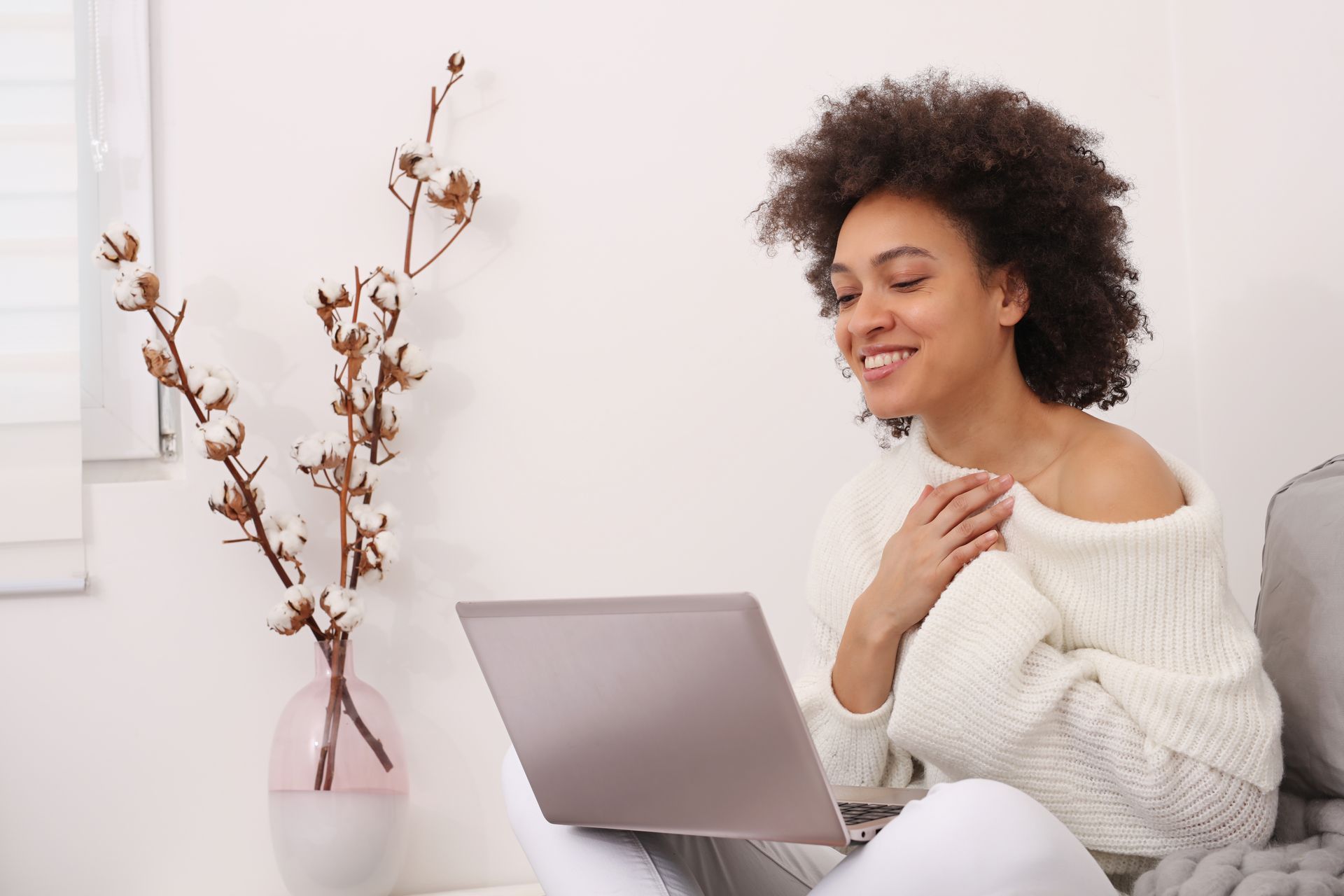 Person smiling beside a laptop on a couch, with a vase of dried branches in a bright room Person smiling beside a laptop on a couch, with a vase of dried branches in a bright room