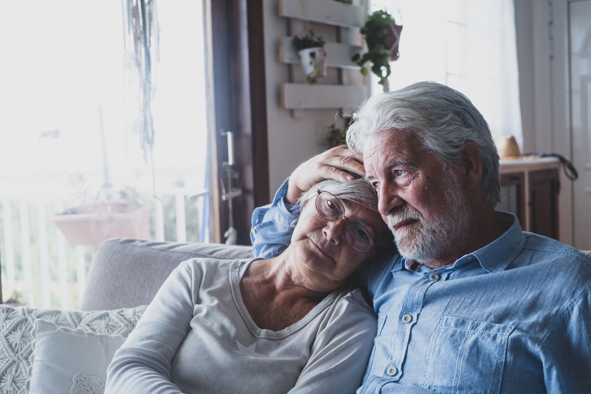 Elderly couple sitting on a couch, with the woman resting her head on the man’s shoulder, both looking calm.