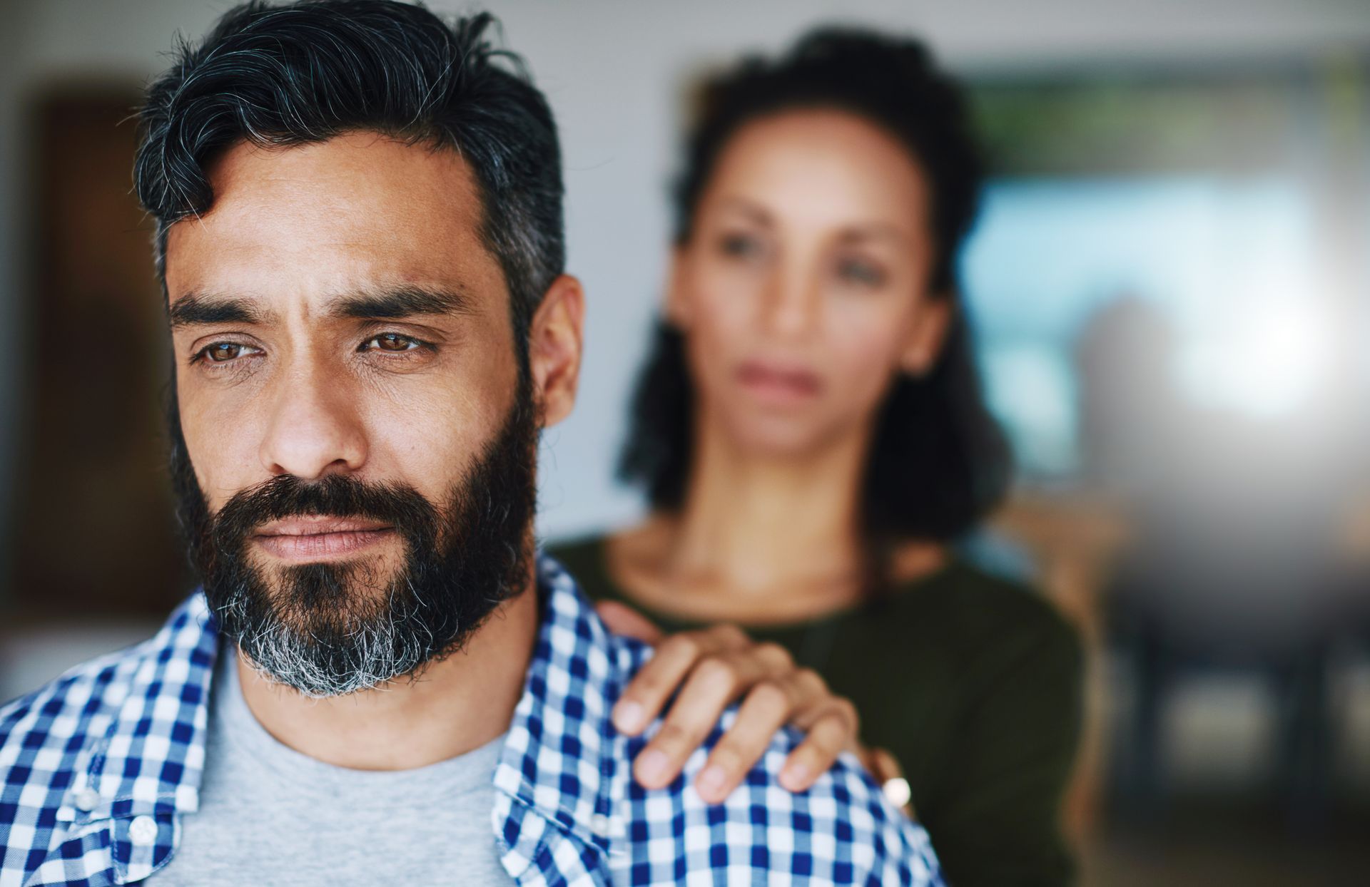 A bearded man in a checkered shirt looks down while a woman stands behind him with a hand on his shoulder.