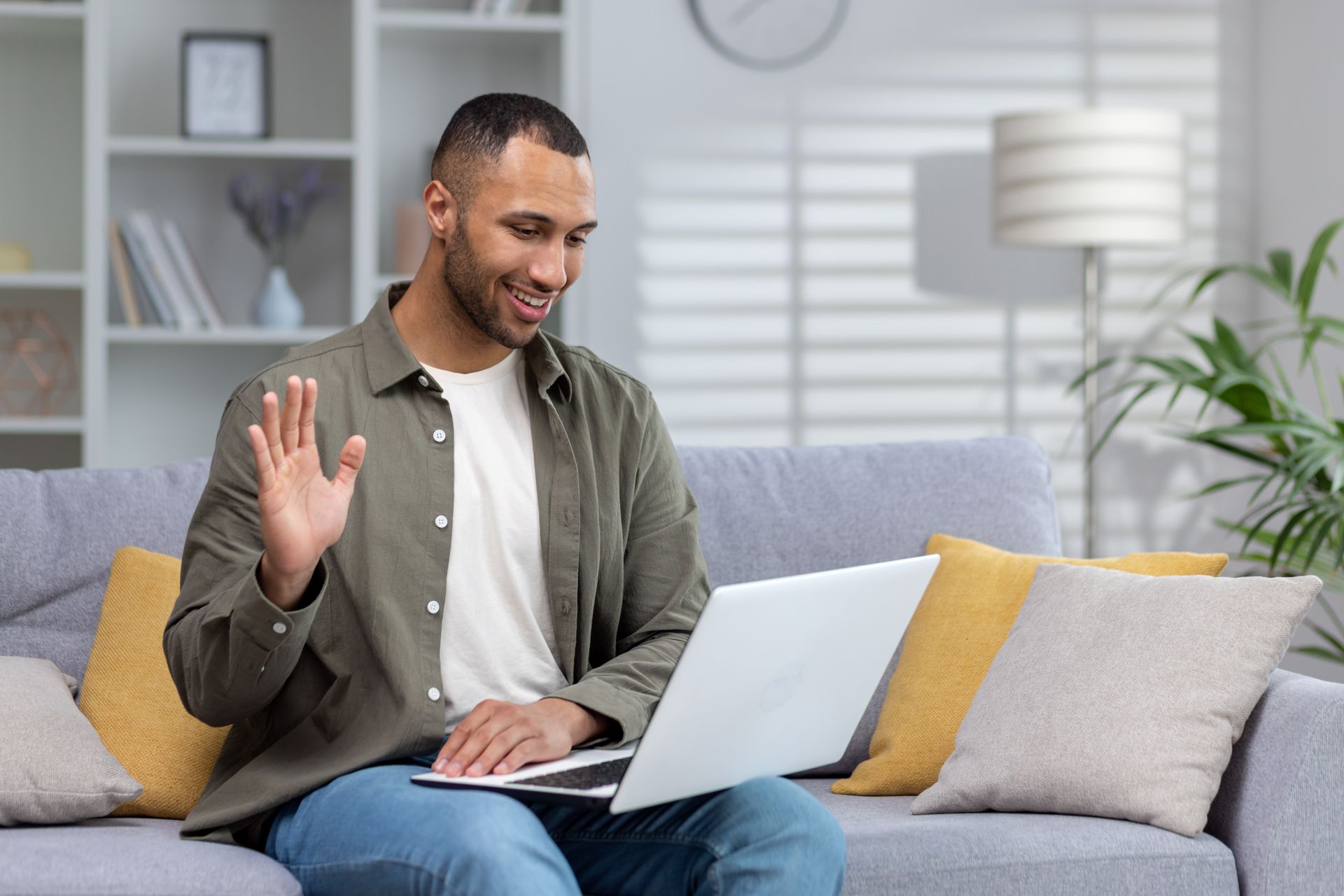 Man waving during a laptop video call on a gray sofa in a bright living room Man waving during a laptop video call on a gray sofa in a bright living room