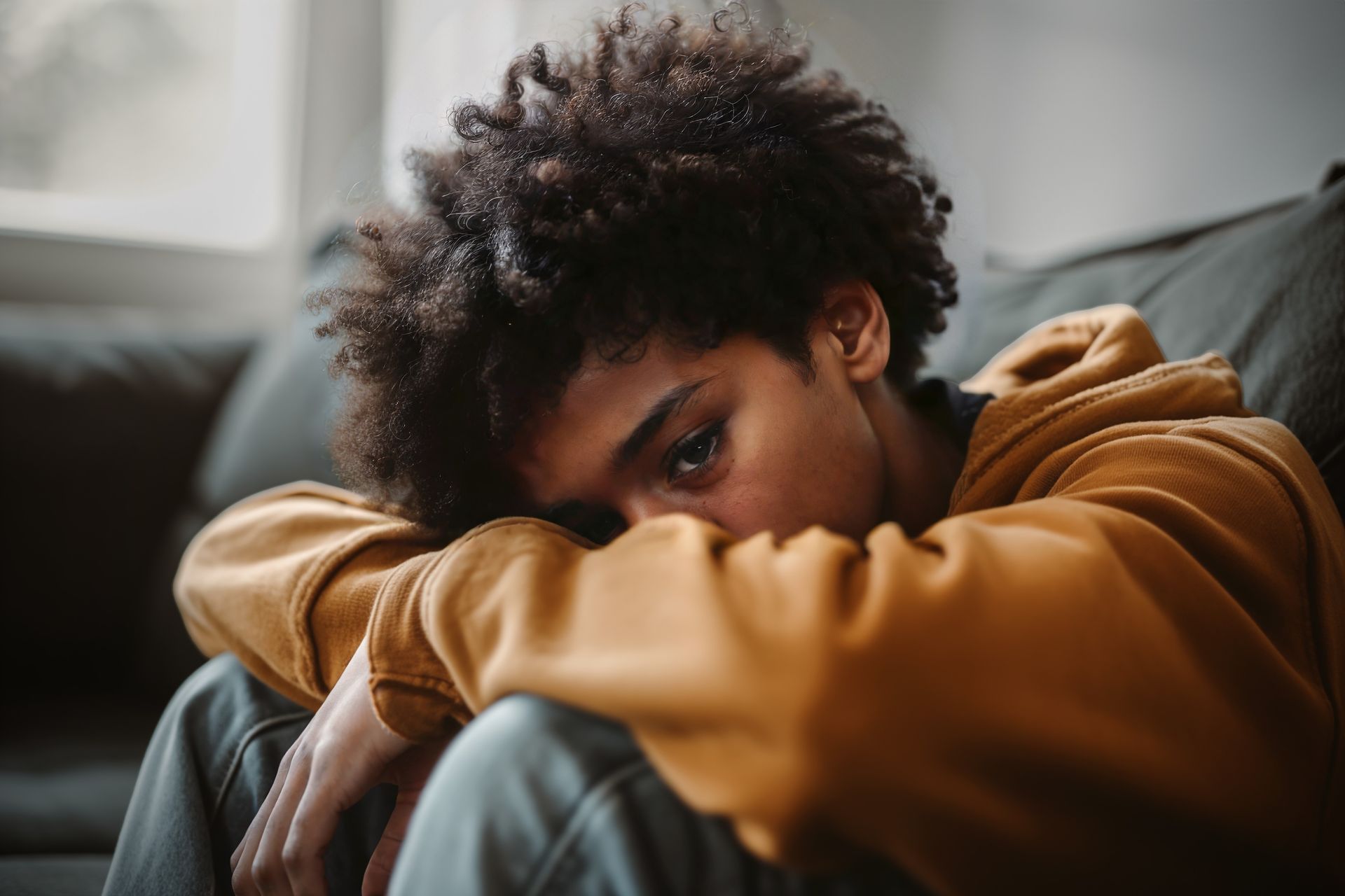Therapist talking with a client on a couch in a bright living room office Person in a mustard hoodie resting their head on folded arms on a couch, looking down sadly.