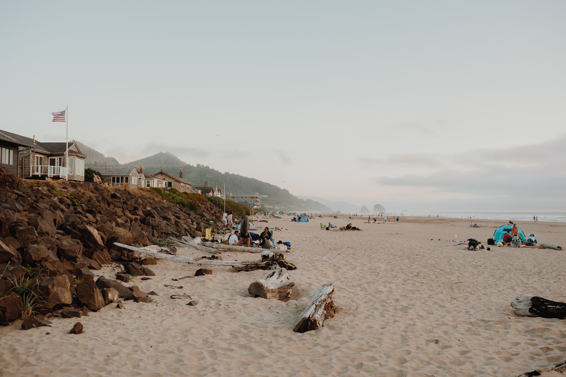 Wide sandy beach with rocks, distant houses, and a hazy mountain coastline under a pale sky