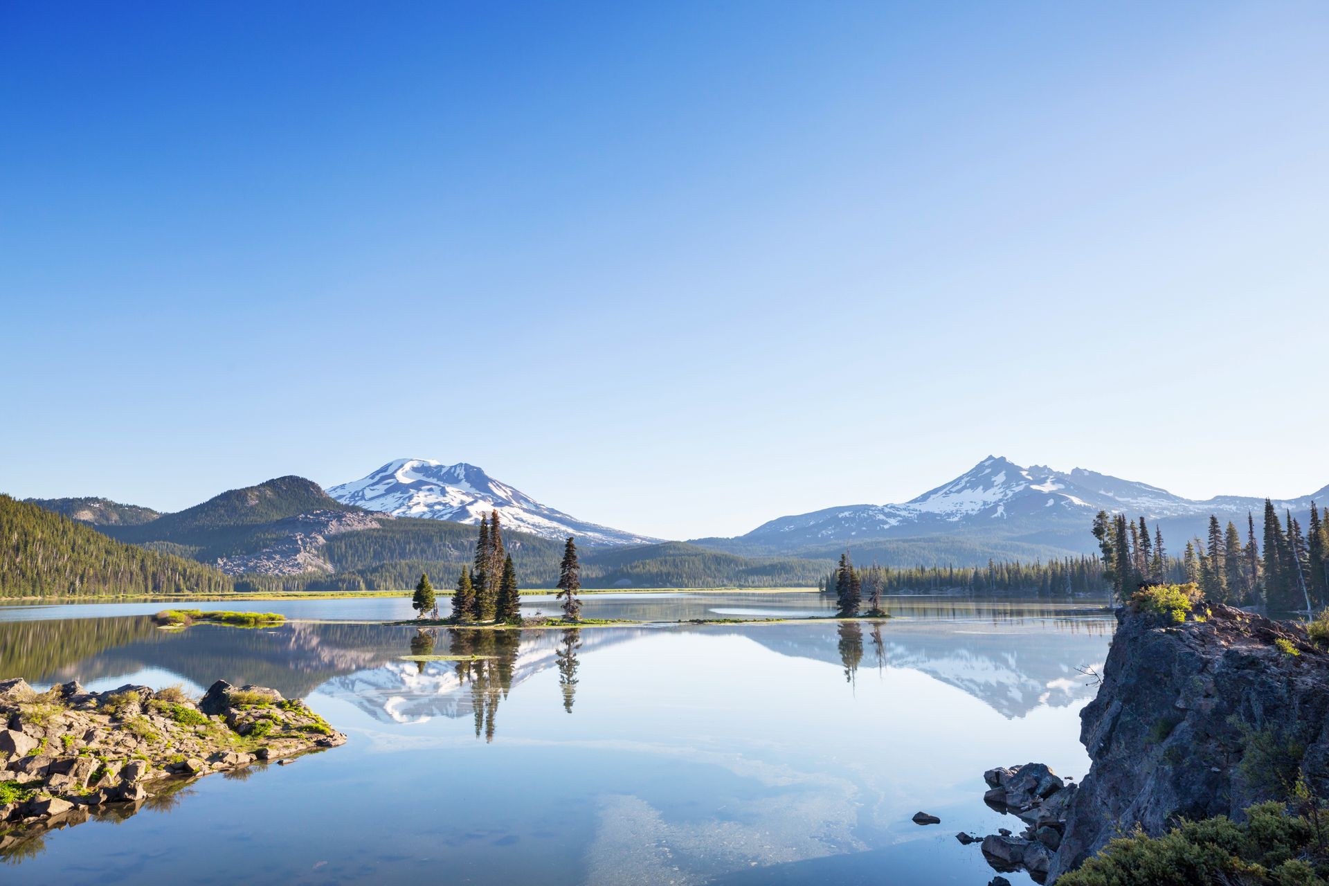 Snowy mountains reflected in a calm lake, framed by rocky shores and evergreens under a clear blue sky