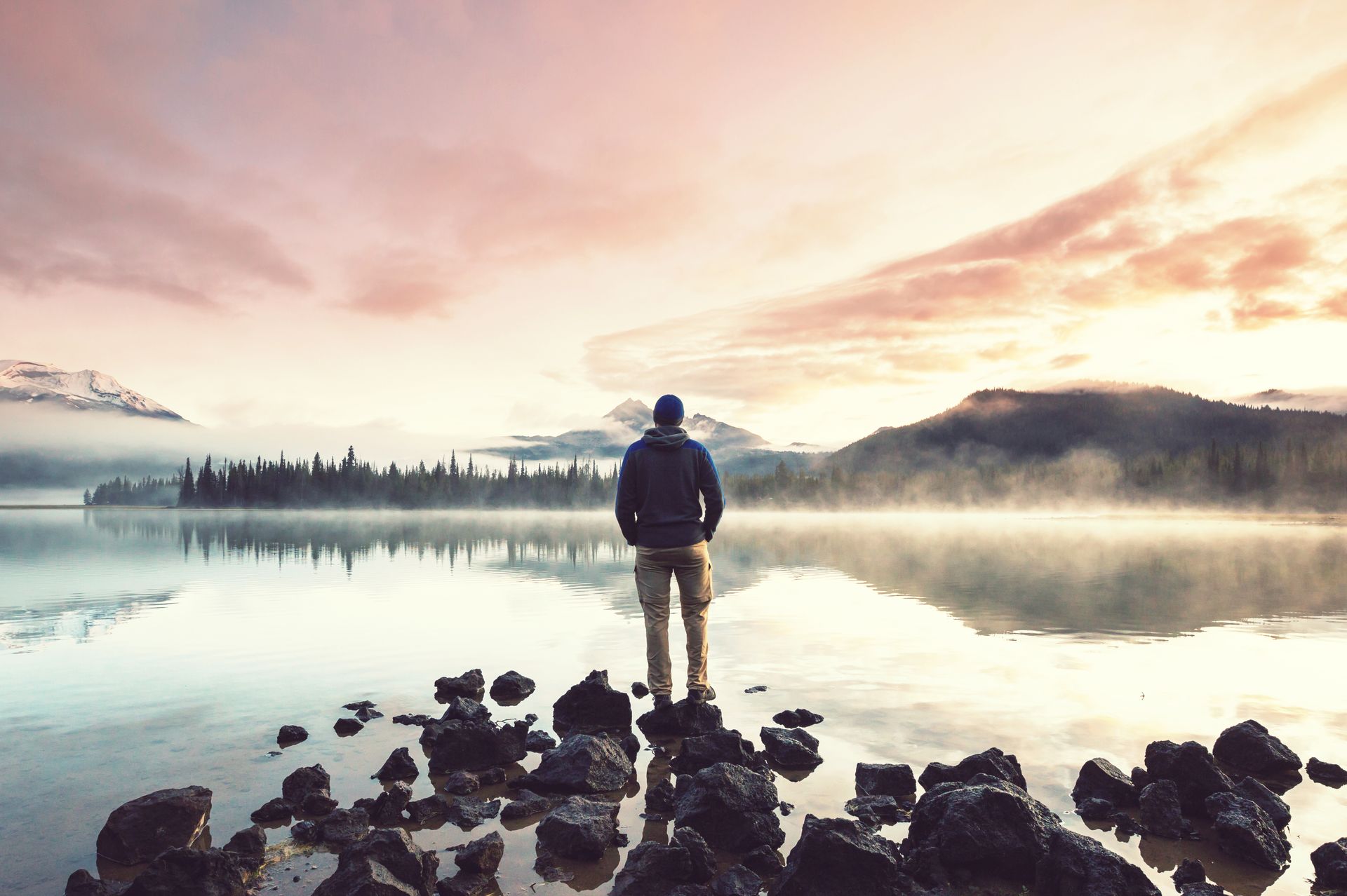 Person stands on rocky shore facing a misty lake at sunrise, with pink clouds and distant trees.