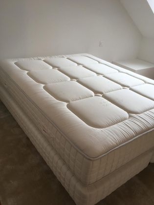 Un matelas de couleur neutre posé sur un sommier dans une chambre peu meublée avec un plafond mansardé.