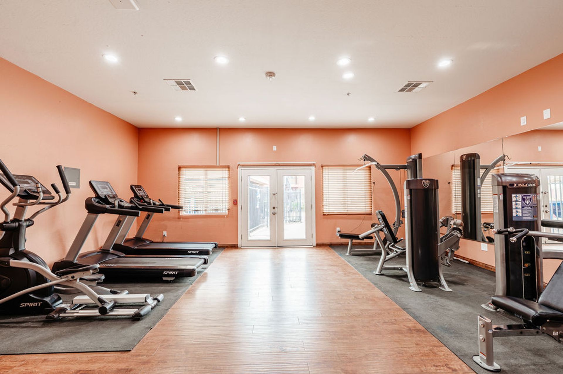 Gym with cardio and weight machines; peach walls, wooden floor, and glass doors.