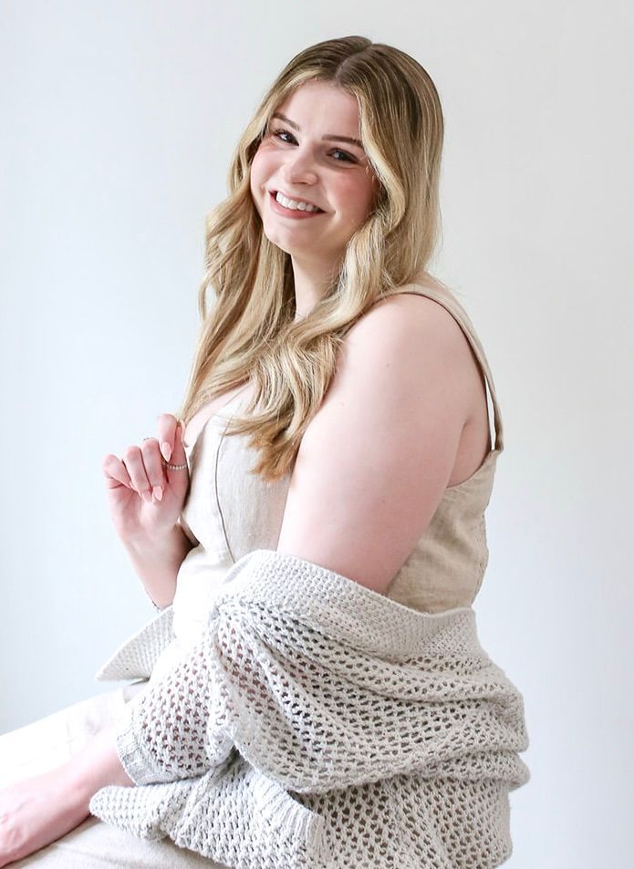 Smiling person with long blonde hair, wearing a beige tank top and a light-colored, open-knit cardigan against a wall.