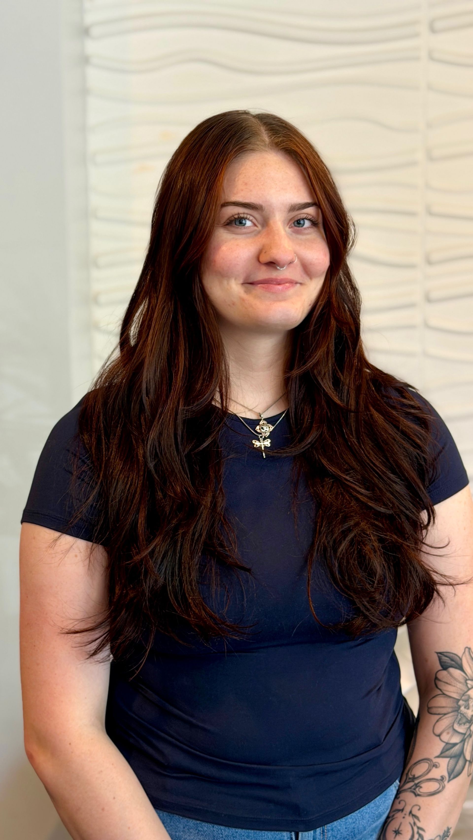 Woman with long dark hair in a navy shirt standing against a light textured wall, smiling gently.
