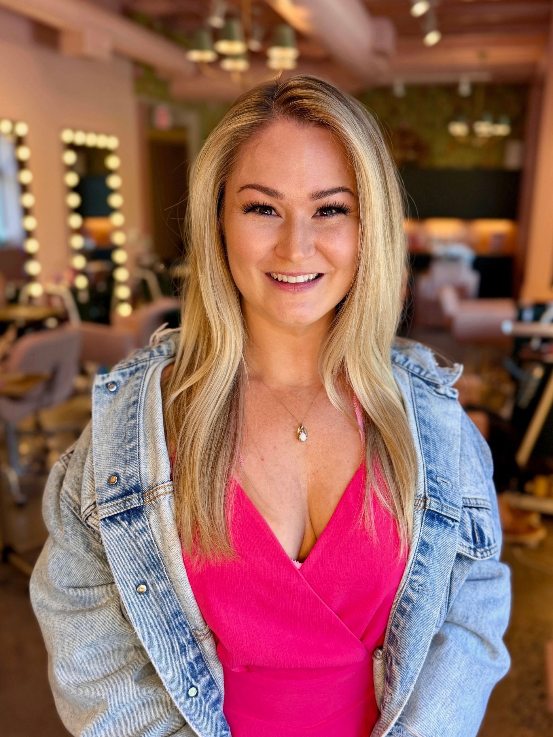 Smiling person in a bright pink top and denim jacket indoors, with vanity lights in the background