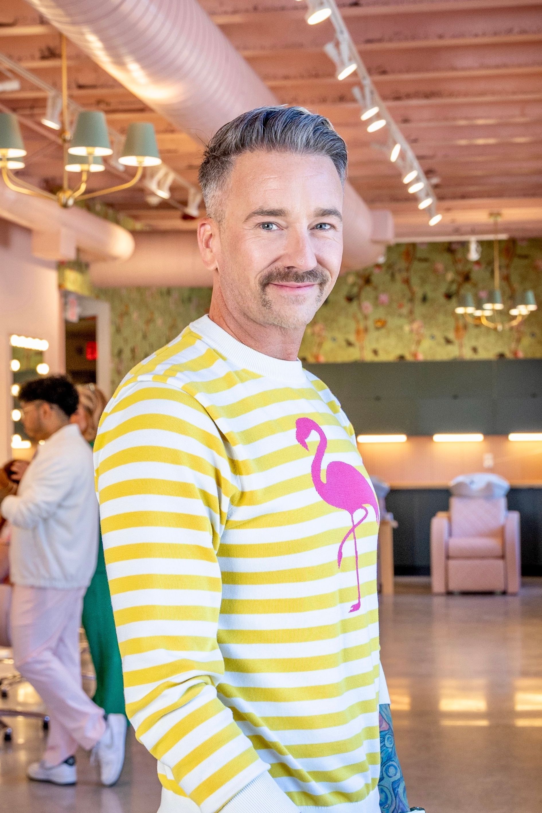 Nate Fox of Luxe Salon & Spa wearing a yellow and white stripe sweater with a flamingo on it and is smiling.