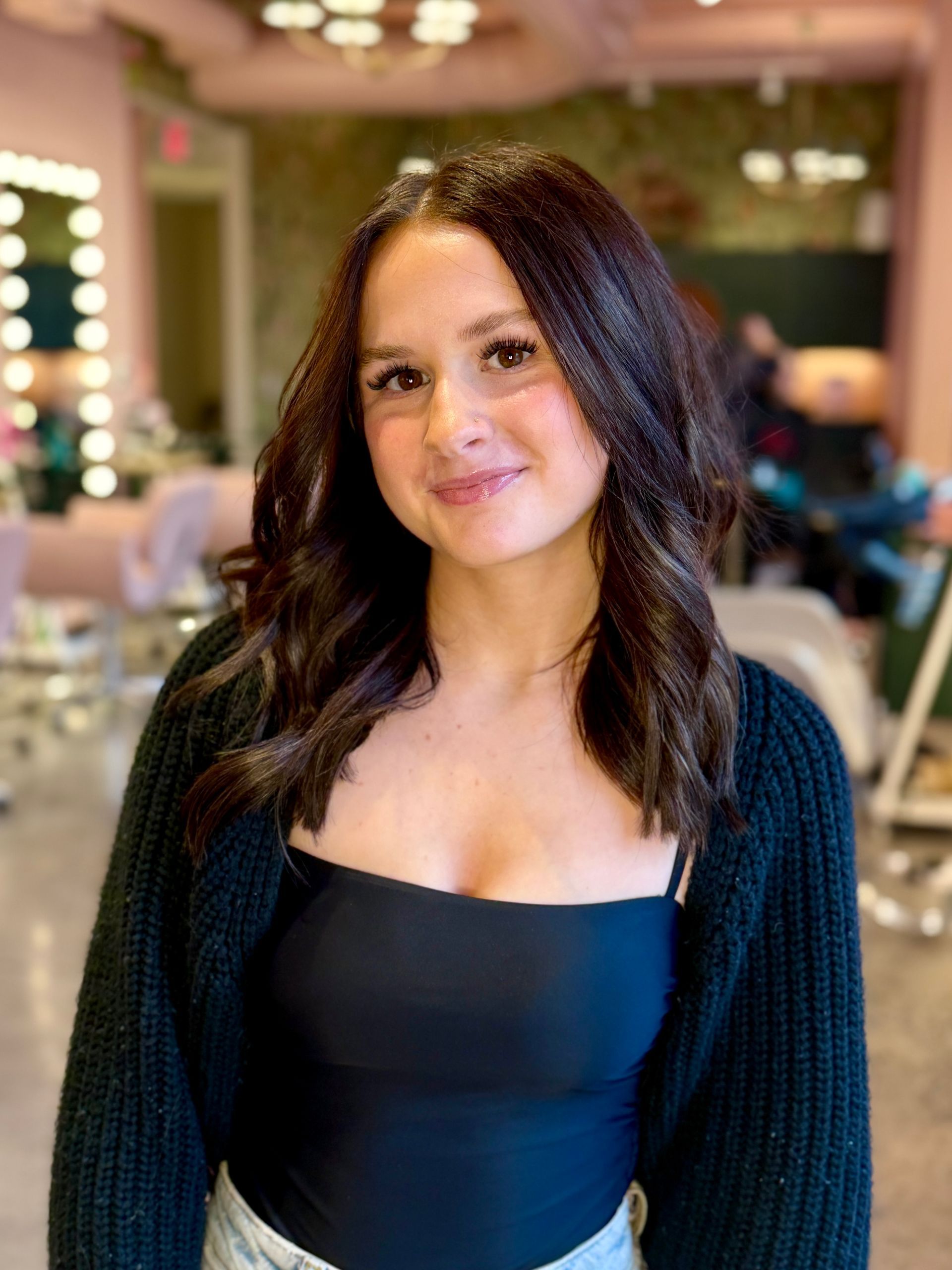 A person with long, dark, wavy hair smiles in a salon with vanity mirror lighting in the background.